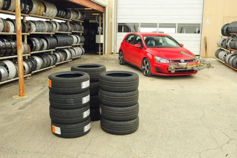 Czerwony Volkswagen Golf GTI z zamontowanym sprzętem do testu opon letnich, obok stosy nowych opon i regały z felgami.