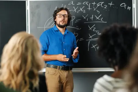 Nauczyciel w niebieskiej koszuli tłumaczy matematykę przy tablicy. Czy nauczyciel jest funkcjonariuszem publicznym? To pytanie nurtuje wielu.