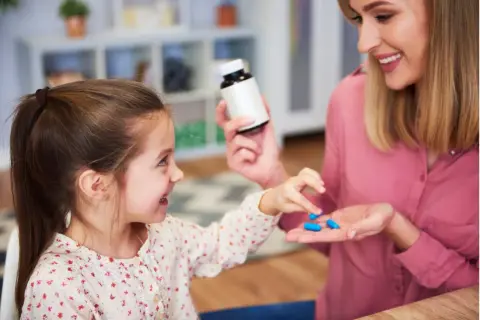 Uśmiechnięta mama podaje dziecku niebieskie kapsułki. Sprawdź, jak podać dziecku antybiotyk, żeby nie wypluło.