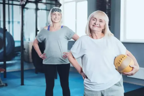 Dwie starsze kobiety ćwiczą na siłowni. Jedna trzyma piłkę. Może to być związane z tabletkami na menopauzę po których się chudnie.