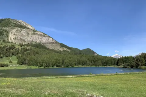 Le lac de Barbeyroux, paisible, reflète le ciel bleu. Des montagnes rocheuses et une forêt dense bordent ses rives verdoyantes.