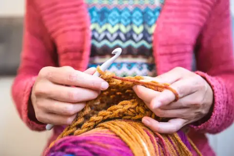Mani che lavorano a maglia con un uncinetto, filato colorato e un maglione rosa. Nonostante sembri rilassante, lavorare a maglia fa male al cuore se si pensa troppo.