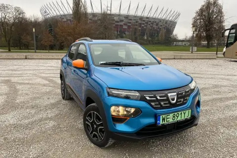 Niebieska Dacia Spring z pomarańczowymi akcentami na tle Stadionu Narodowego. Dacia Spring opinie – miejski elektryk gotowy na przygodę.
