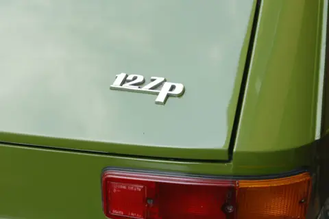 Chromowana plakietka "127p" na zielonej karoserii klasycznego Fiata 127. Widoczny fragment tylnej lampy.