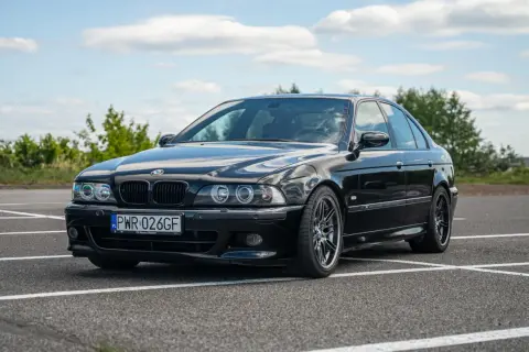 Czarne BMW E39 na parkingu, z widocznym przodem i bokiem samochodu.