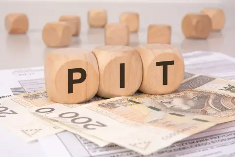 Drewniane klocki z literami "PIT" leżą na banknotach 200 zł i dokumentach podatkowych. To symboliczne przedstawienie podatku od darowizny.