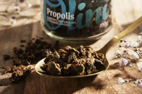 Łyżka pełna propolisu, ukazująca jego naturalne, zbite grudki. Słoik z napisem "Propolis" w tle podkreśla jego cenne właściwości.