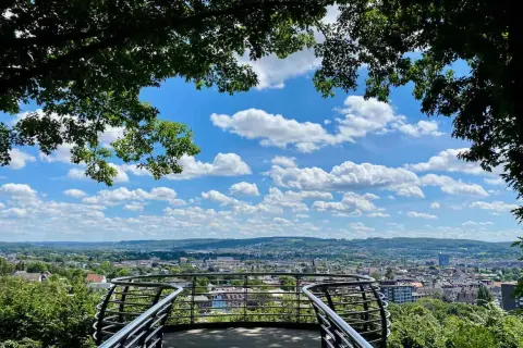 Blick vom Aussichtspunkt Wuppertal über die Stadt. Ein geschwungenes Geländer rahmt die weite Sicht auf Häuser und grüne Hügel unter einem blauen Himmel mit weißen Wolken ein.