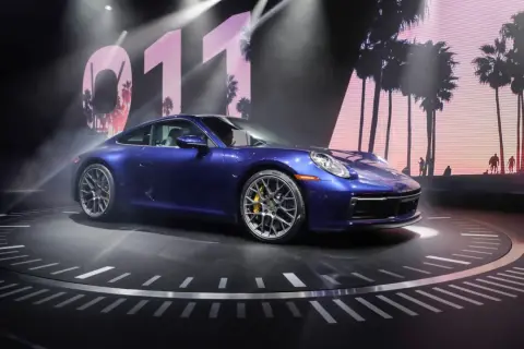 Un Porsche 911 992 azul metálico, con pinzas de freno amarillas, se exhibe sobre un escenario circular con un fondo de palmeras y el número 011.