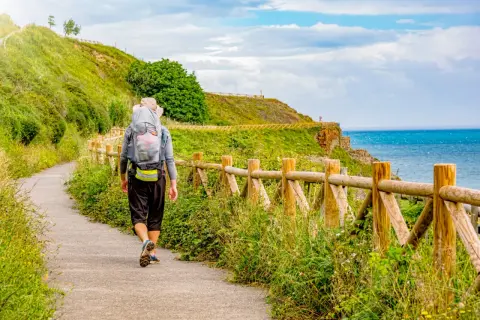 Vandrare på el camino de Santiago längs en kuststig med havet till höger och en grön kulle till vänster.
