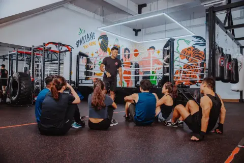 A boxing coach instructs a group sitting on the floor, preparing them for boxing reflex training. The gym features a boxing ring and punching bags.