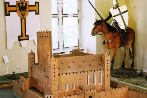 Ein deutscher Orden Ritter auf einem Pferd, mit Lanze und Schild, steht vor einem Modell einer mittelalterlichen Burg.