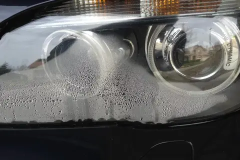 Zaparowane reflektory BMW z licznymi kropelkami wody wewnątrz. Widać odbicie domu i drzew.