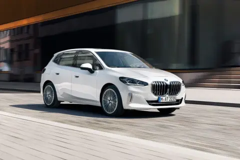 Białe BMW 218i sunie po miejskiej ulicy, odbijając światło od nowoczesnej fasady budynku.