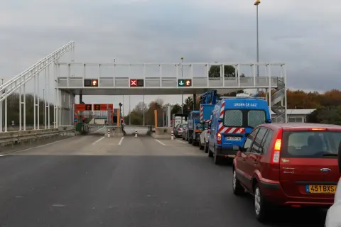 Voiture rouge et camions bleus dans une file d'attente au péage flux libre A13. Lumières rouges et vertes sur les panneaux.
