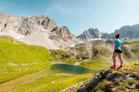 Une femme admire un lac de montagne lors d'une randonnée à Courchevel. Paysage alpin spectaculaire avec des montagnes rocheuses et des prairies verdoyantes.
