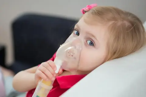 Mała dziewczynka z niebieskimi oczami korzysta z nebulizatora, co jest częścią leczenia mukowiscydozy.