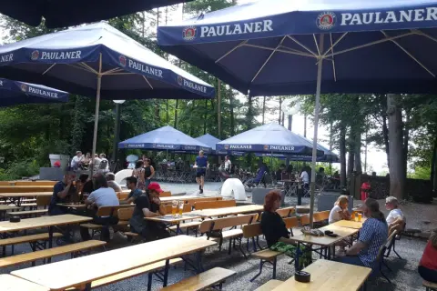 Ein sonniger Tag im Paulaner Biergarten. Gäste sitzen an Holztischen unter großen Sonnenschirmen und genießen die Atmosphäre.
