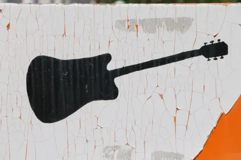 Czarna sylwetka gitary elektrycznej na białym, popękanym tle. Wyobraź sobie rockowy koncert i wirtuoza grającego na tym instrumencie.