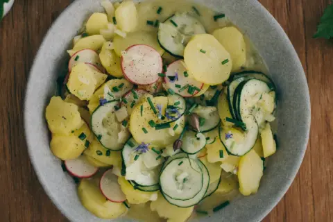 Kartoffelsalat mit Gurken, Radieschen und Schnittlauch, garniert mit kleinen blauen Blüten. Ein frischer Genuss auf Holztisch.