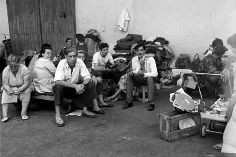 Un groupe de personnes, dont des hommes, des femmes et un bébé, attendent, reflétant le caractère des pieds-noirs dans l'attente.