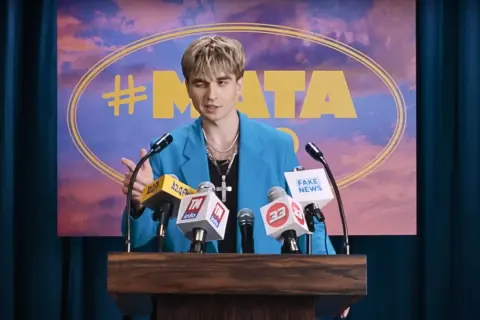 Mata w niebieskiej marynarce przemawia za mównicą z mikrofonami, na tle napisu "#MATA".