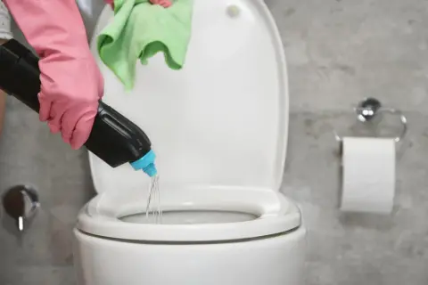 Mano con guante rosa vierte agua fuerte para que sirve en el inodoro. Paño verde en mano.