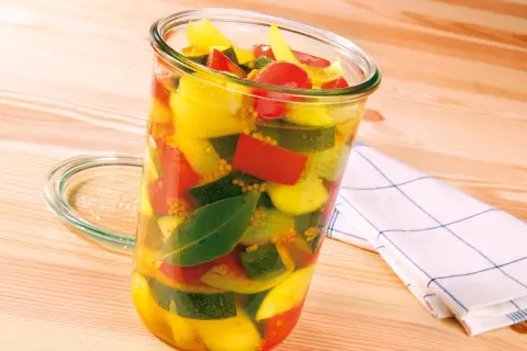 Ein Glas mit bunter Mischung aus Zucchini, Paprika und Tomaten, bereit zum Zucchini einkochen. Senfkörner und Lorbeerblätter sind ebenfalls sichtbar.