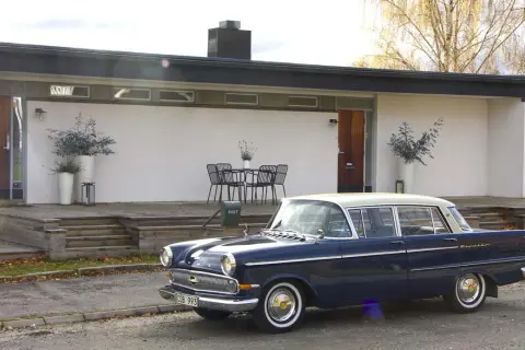 En mörkblå Opel Kapitän från 1960 står parkerad framför ett modernt hus med träaltan.