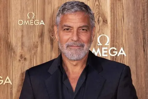 Un homme avec des cheveux gris courts et une barbe, portant un costume sombre, pose devant un mur en bois avec le logo Omega.