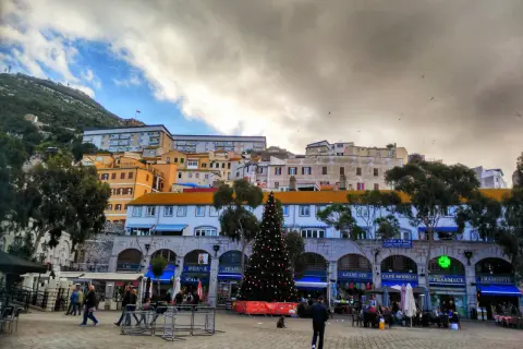 Árbol de Navidad en la plaza de Gibraltar, con edificios coloridos y el Peñón al fondo. Un día nublado, pero festivo.