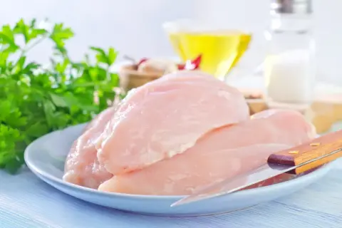 Surowe piersi kurczaka na talerzu z natką pietruszki, oliwą i solniczką w tle.