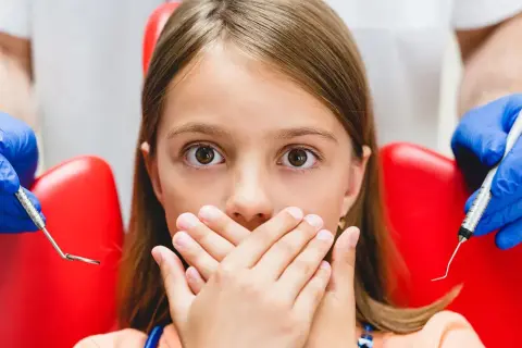 Przerażona dziewczynka zakrywa usta dłońmi, gdy dentysta przygotowuje się do podania znieczulenia. Wokół niej widać czerwony fotel dentystyczny i narzędzia.