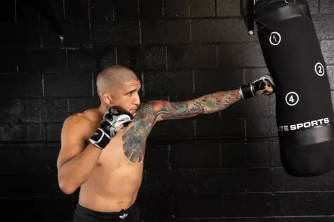 A tattooed man in boxing gloves performs a punching bag workout, his fist connecting with the bag.