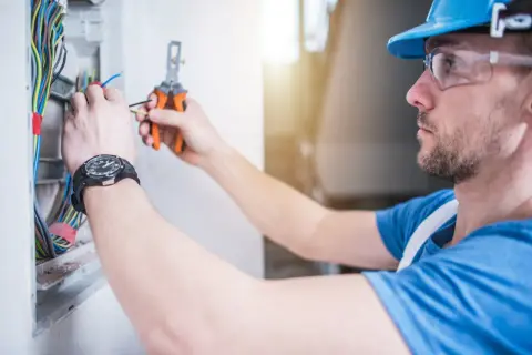 Elektryk przemysłowy w kasku i okularach ochronnych pracuje przy skrzynce z przewodami, używając obcinaczek.