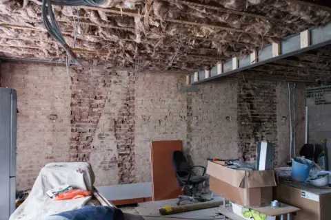 Pågår renovering av funkishus. Synligt bjälklag med isolering och tegelväggar. Material och verktyg ligger utspridda.
