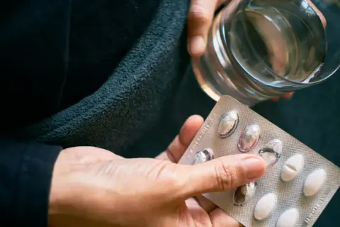 Dłoń trzyma blister z tabletkami i szklankę wody. Czy to próba zwalczenia nadmiaru witaminy B12?