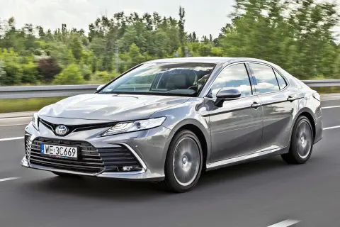 Srebrna Toyota Camry 2023 jedzie po drodze, otoczona zielenią drzew.