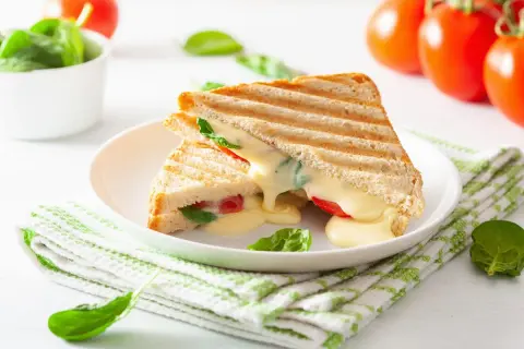 Grillowany tost z serem, pomidorami i szpinakiem na białym talerzu, obok pomidory i miseczka szpinaku.