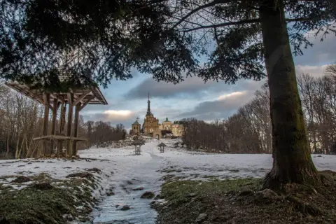 Zimowe szlaki w Górach Świętokrzyskich prowadzą do Świętej Katarzyny. Na pierwszym planie drewniana wiata z krzyżem, w oddali klasztor.