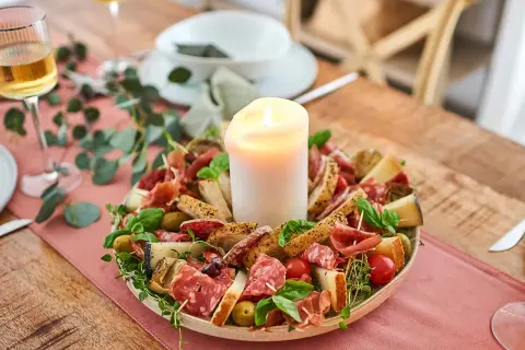 Antipasti selber machen: Eine festliche Platte mit Wurst, Käse, Brot, Oliven und Tomaten, umgeben von grünen Zweigen, mit einer Kerze in der Mitte.
