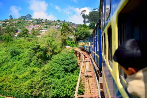 Ett blått och gult tåg i Indien färdas längs en bergssluttning täckt av frodig grönska och färgglada hus.