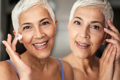Zwei Frauen mit kurzen weißen Haaren lächeln. Eine trägt Creme auf ihre Wange auf, um trockene Haut im Alter zu bekämpfen.
