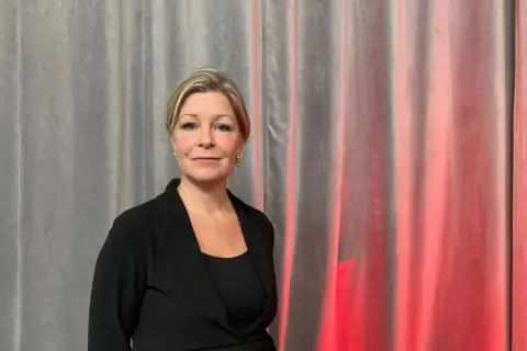 Johanna Fager Wettergren, en kvinna med blont hår och blå ögon, ler mot kameran framför en grå ridå med röd belysning.