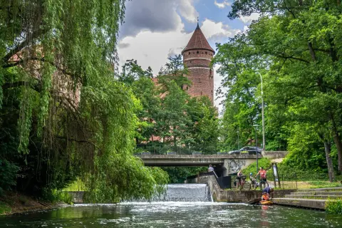 Olsztyn atrakcje dla dorosłych: malowniczy widok na zamek nad rzeką, z mostem i ludźmi odpoczywającymi nad wodą.