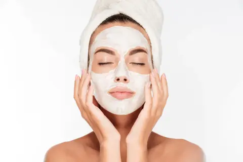 Une femme avec une serviette sur la tête applique un masque blanc sur son visage, espérant atténuer les marques de masque cicatrice acné.