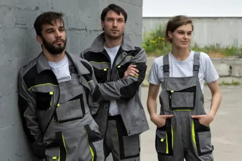 Trzech pracowników w szarych kombinezonach z neonowymi akcentami. Polski producent odzieży roboczej prezentuje nowoczesne ubrania.