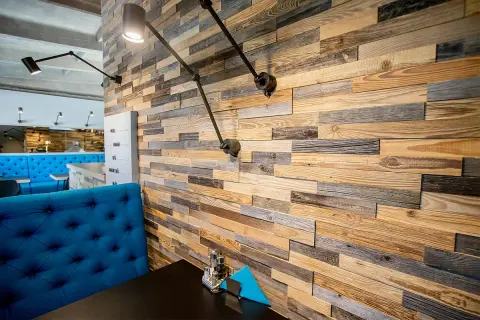 Mur en bois brut, aux teintes variées, évoquant l'essence de bois authentique. Des appliques industrielles éclairent ce décor chaleureux.