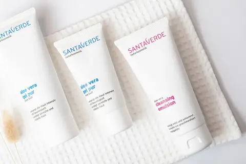 Drei weiße Tuben mit Naturkosmetik, darunter Produkte mit reiner Aloe Vera, liegen auf einem weißen Handtuch.