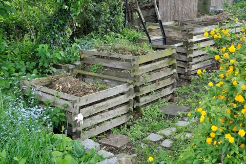 Holzkomposter im Garten, bereit, um kompost anlegen zu beginnen. Gelbe und blaue Blumen blühen daneben.